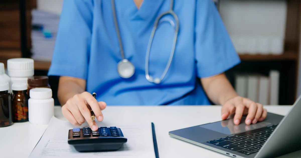 Médico calculando custos com calculadora e laptop em mesa de consultório - cálculo do Fator R para clínica médica PJ